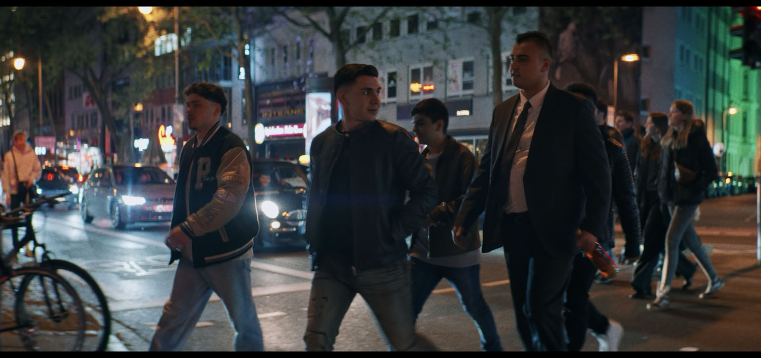 A group of people walking on a city street at night, with cars and bicycles nearby, and buildings illuminated in the background.